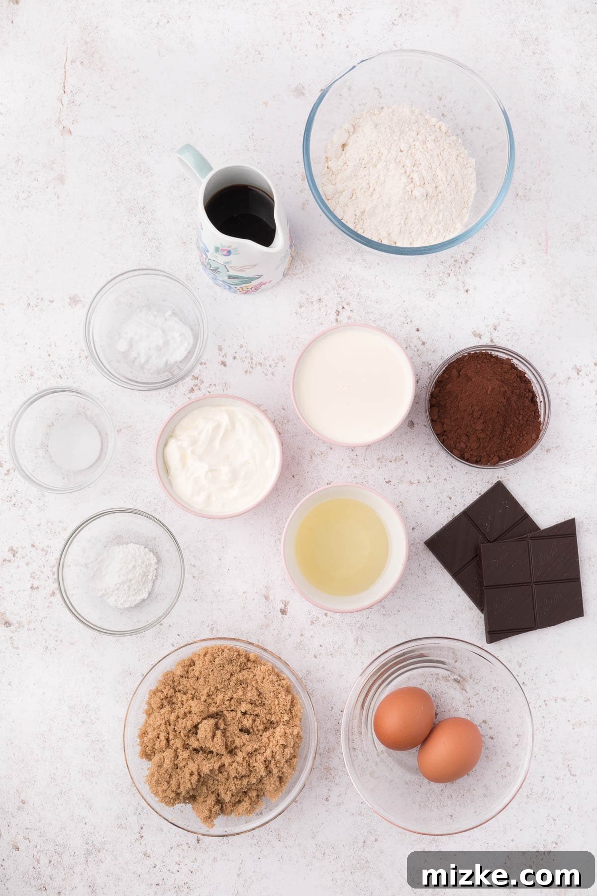 Assorted ingredients for mini bundt cakes, including flour, cocoa powder, sugar, eggs, and cream, neatly arranged on a kitchen counter.