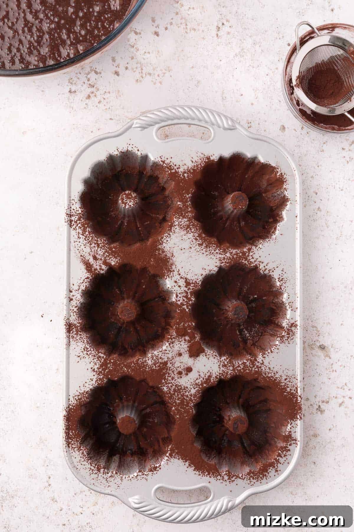 A metal mini bundt pan with multiple cavities, each meticulously dusted with cocoa powder, ready for batter.