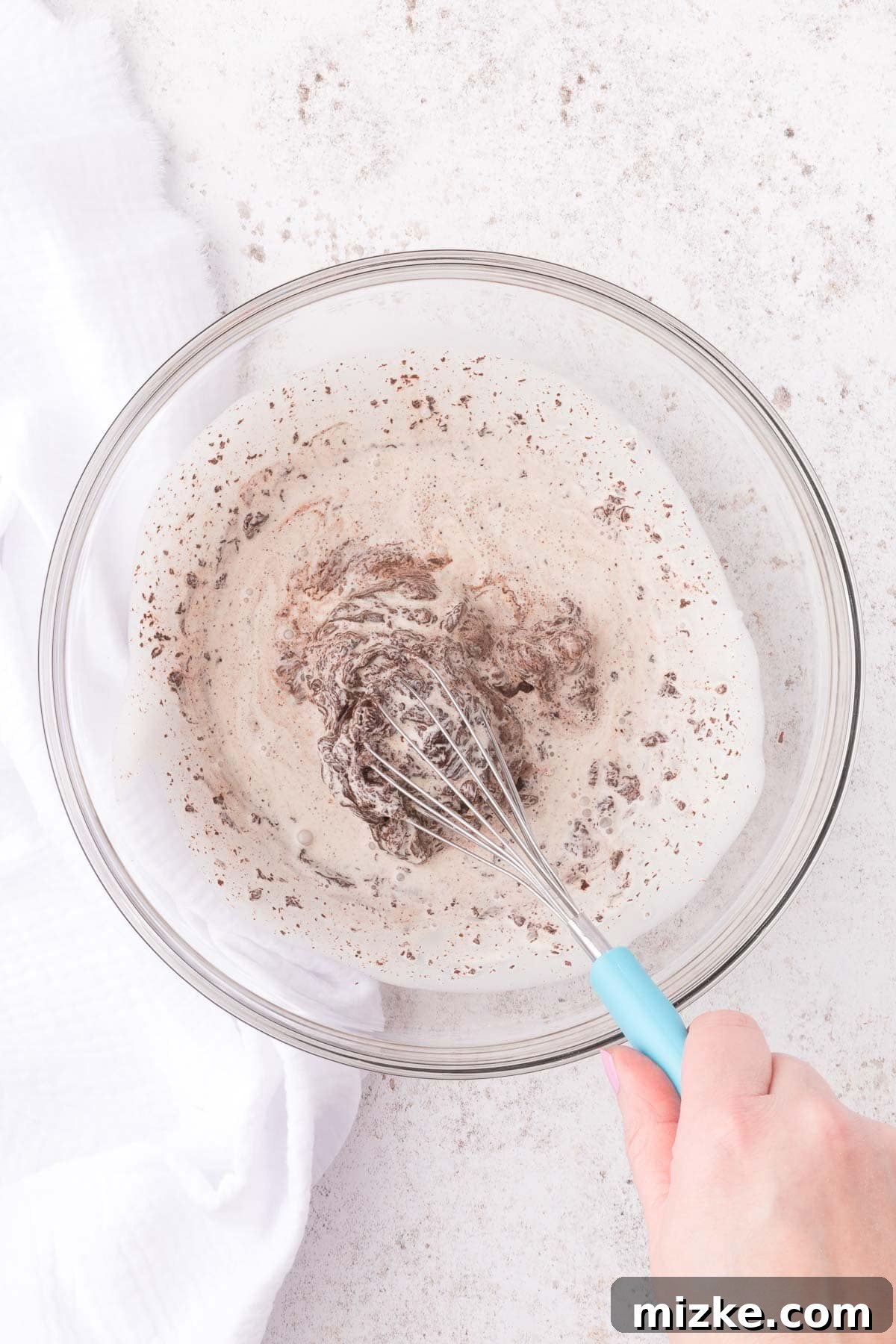 A close-up view of warm heavy cream being poured over finely chopped semi-sweet chocolate in a clear glass bowl, beginning the ganache process.