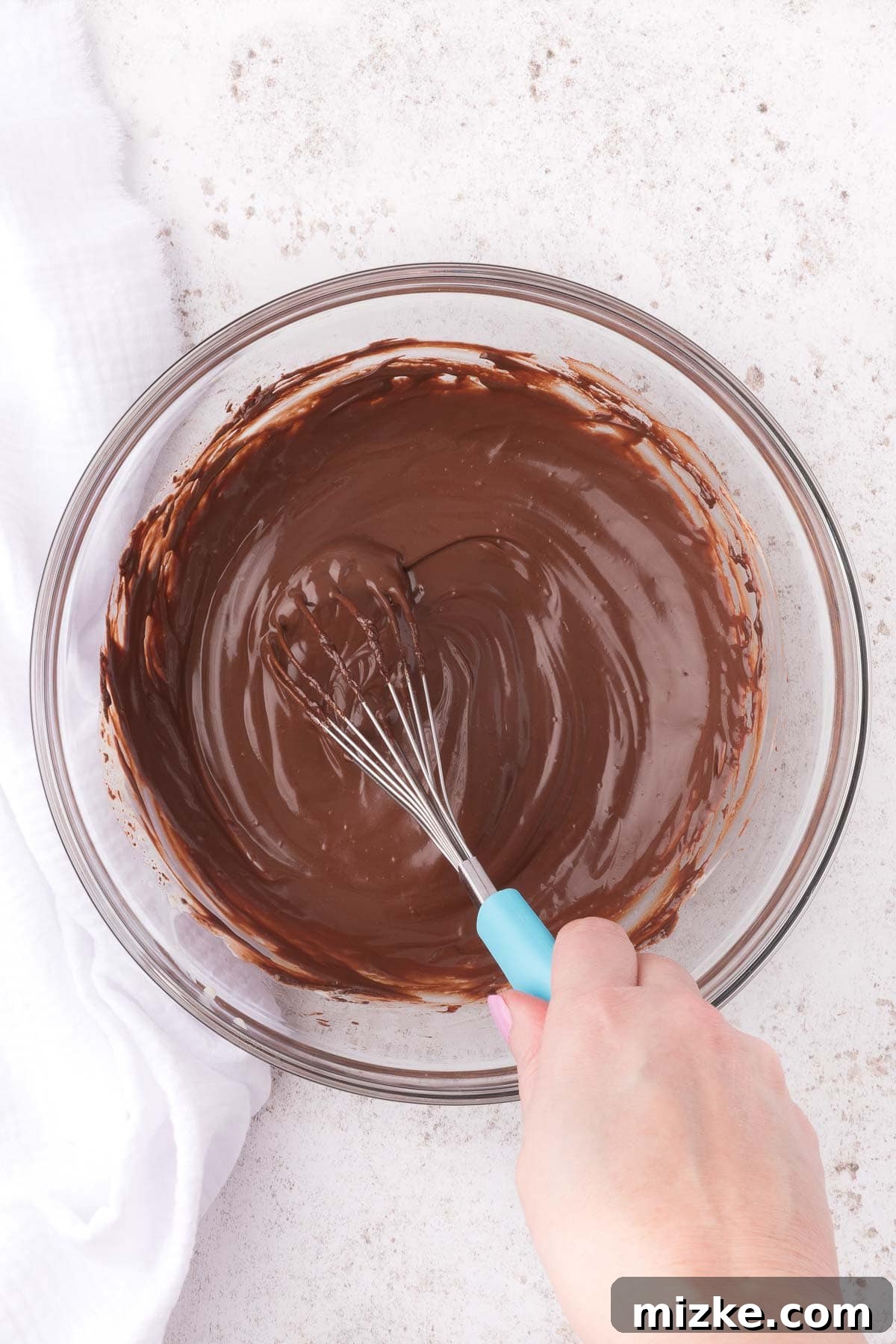 A whisk smoothly blending warm cream and melted chocolate in a glass bowl, creating a rich, glossy chocolate ganache.