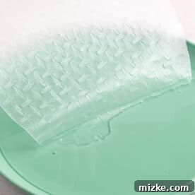 A round rice paper wrapper being gently dipped into a shallow bowl of warm water on a teal plate, softening it for rolling.