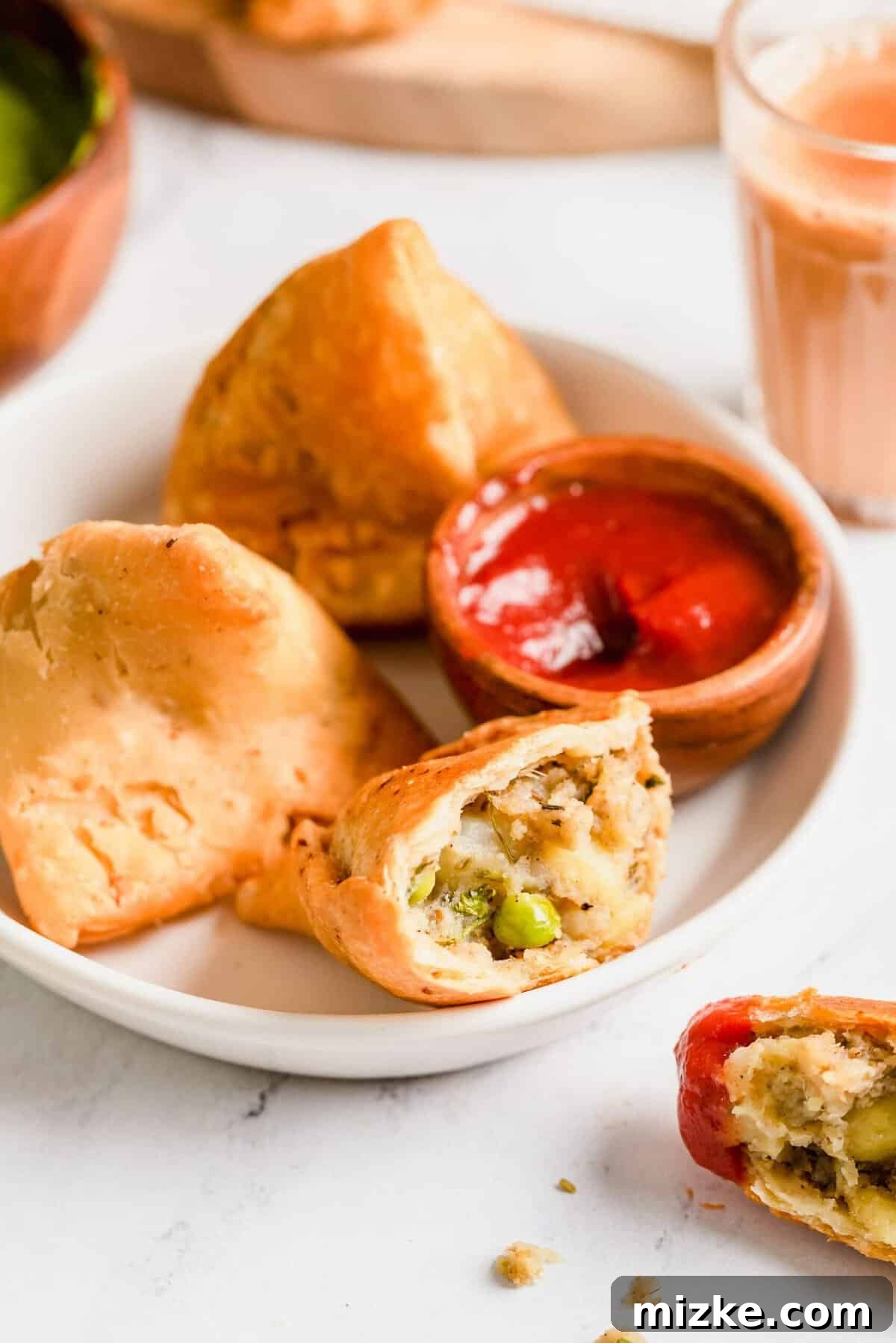 Golden Crispy Vegetable Samosas 4 A plate of golden-brown vegetable samosas with a small bowl of green dipping sauce. One samosa is cut open to reveal the flavorful potato and pea filling.