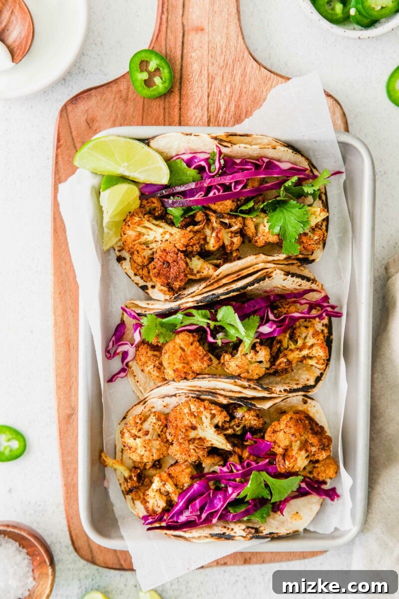 Crispy Spiced Vegan Cauliflower Tacos 2 Three golden-brown roasted cauliflower tacos on a metal baking sheet lined with parchment paper, garnished with fresh cilantro and a wedge of lime, showcasing their vibrant appeal.