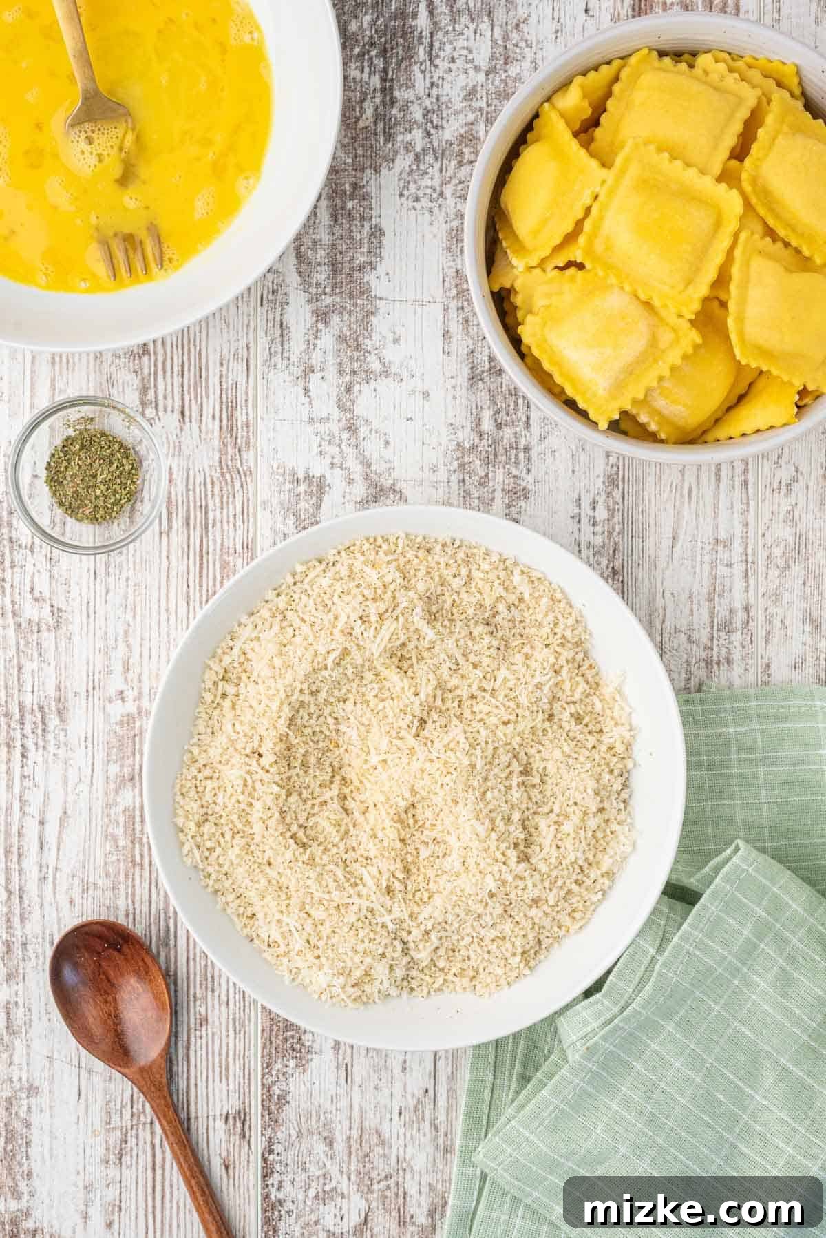 Air Fryer Toasted Ravioli 4 A white shallow plate holds a finely tuned mixture of panko breadcrumbs, grated Parmesan cheese, and various spices, with a wooden spoon positioned beside it for mixing.