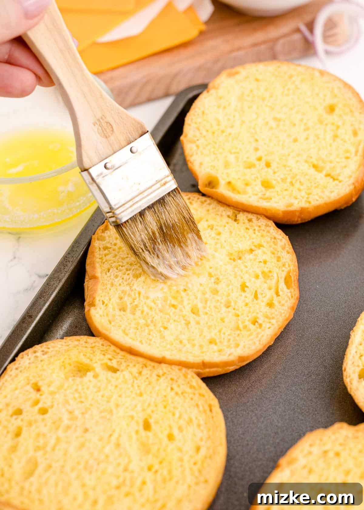 Melted butter being generously brushed onto the cut sides of brioche hamburger buns before toasting.