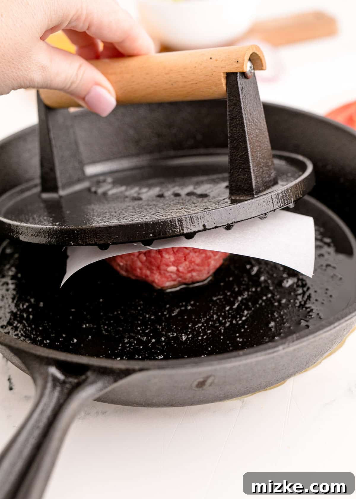 A ball of ground beef being vigorously pressed down onto a hot cast iron skillet using a heavy meat mallet over parchment paper, creating a thin patty with a crust.