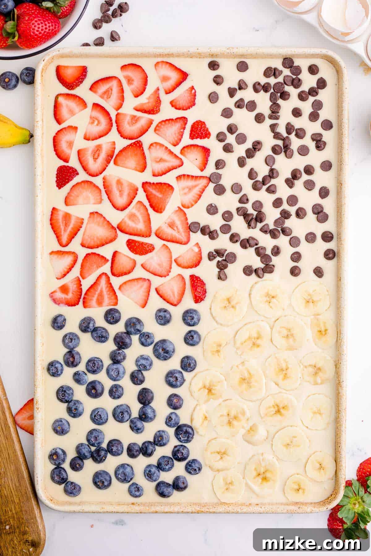 One-Pan Fruity Pancakes 11 A sheet pan filled with pancake batter, neatly divided into four sections, each topped with a different delicious ingredient: strawberries, chocolate chips, blueberries, and sliced bananas.