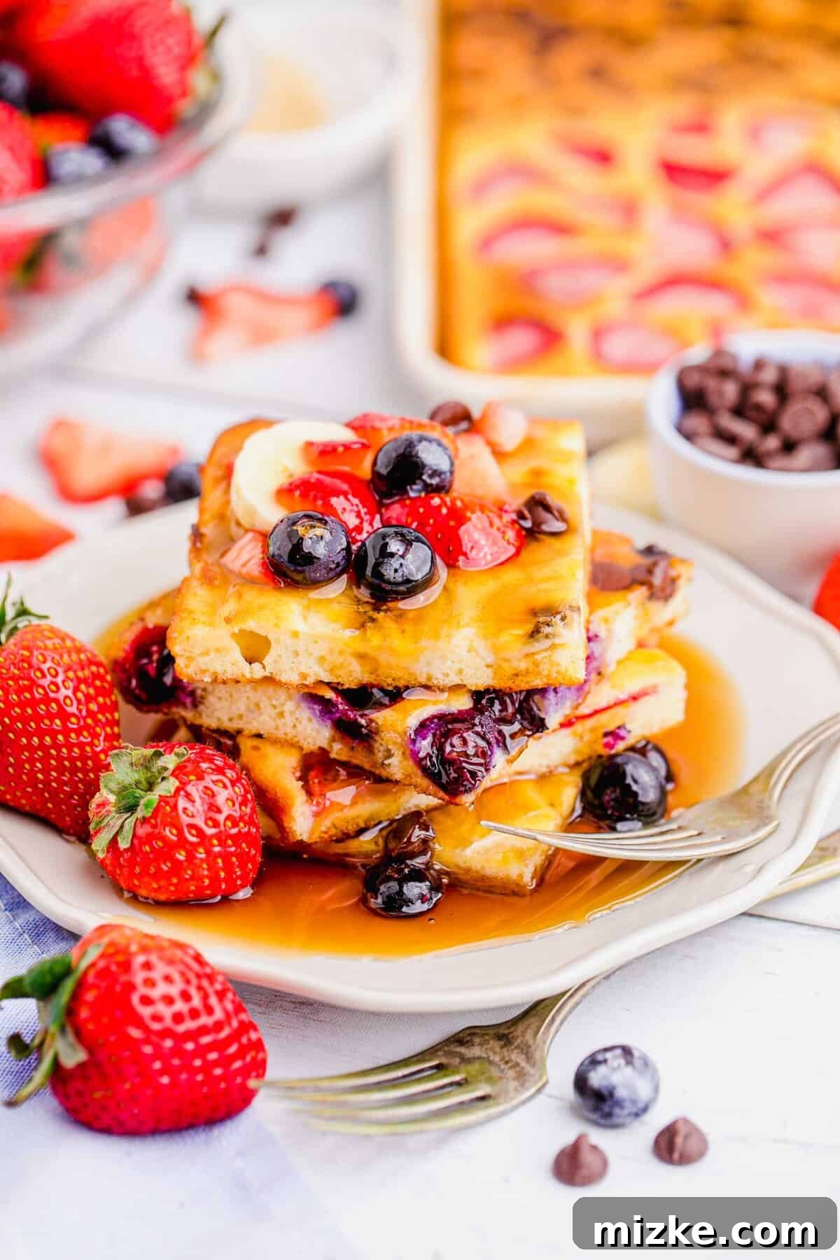 One-Pan Fruity Pancakes 13 A delightful stack of square-cut pancakes, generously topped with fresh strawberries and blueberries, drizzled with golden maple syrup, on a pristine white plate.