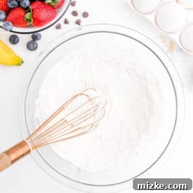 One-Pan Fruity Pancakes 16 A copper whisk is gently stirring flour in a clear mixing bowl, preparing the dry ingredients for the pancake batter.
