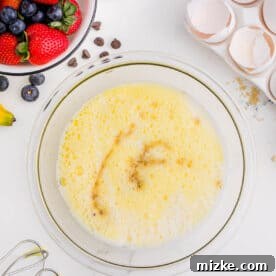 One-Pan Fruity Pancakes 17 A bowl of whisked egg mixture with a splash of vanilla, surrounded by broken eggshells and fresh berries, highlighting the liquid ingredients.