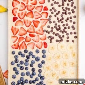 One-Pan Fruity Pancakes 20 A sheet pan filled with pancake batter, neatly divided into four sections, each topped with a different delicious ingredient: strawberries, chocolate chips, blueberries, and sliced bananas.