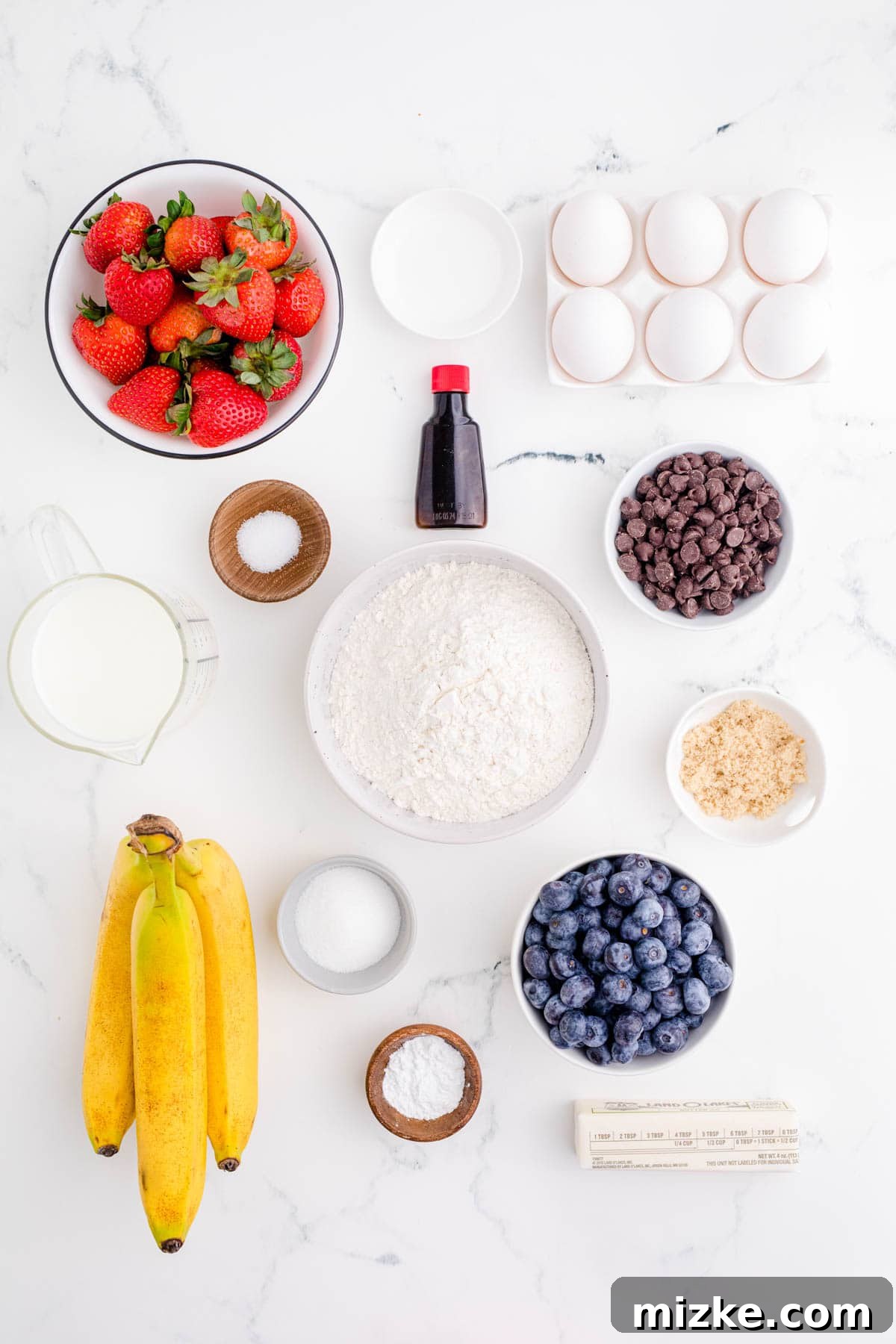 One-Pan Fruity Pancakes 3 A collection of fresh ingredients laid out for making fruit pancakes, including flour, sugar, eggs, milk, blueberries, strawberries, and bananas.