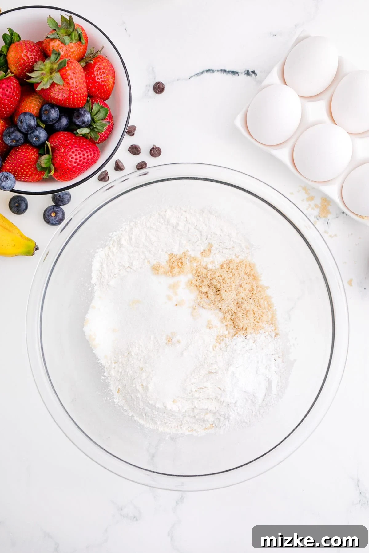 One-Pan Fruity Pancakes 4 Dry pancake ingredients including flour, white sugar, brown sugar, baking powder, and salt in a clear bowl next to fresh eggs and fresh berries, indicating readiness to mix.