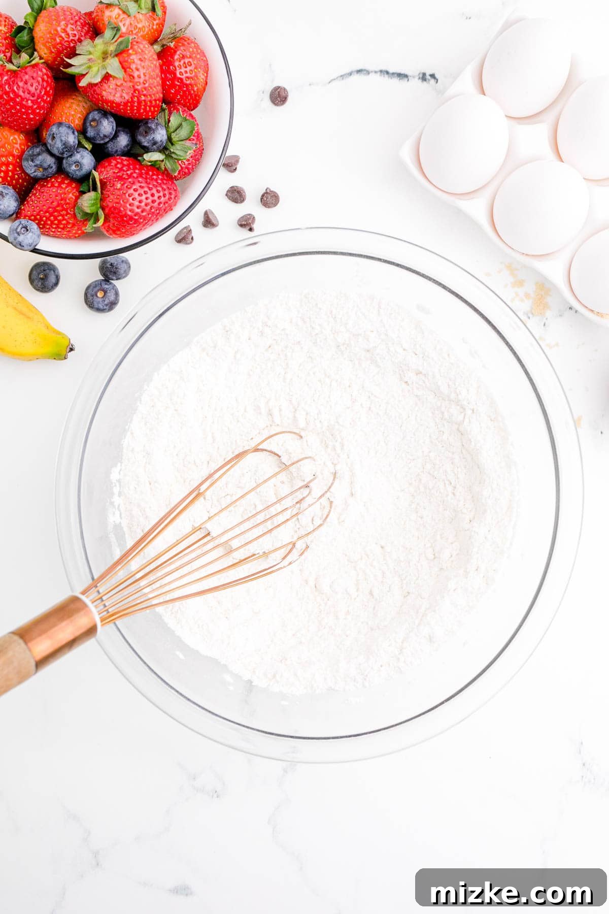 One-Pan Fruity Pancakes 5 A copper whisk is gently stirring flour in a clear mixing bowl, preparing the dry ingredients for the pancake batter.