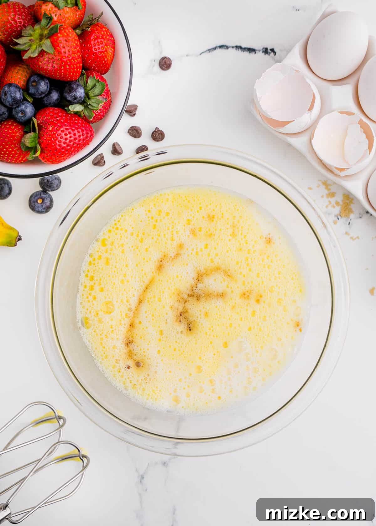 One-Pan Fruity Pancakes 7 A bowl of whisked egg mixture with a splash of vanilla, surrounded by broken eggshells and fresh berries, highlighting the liquid ingredients.