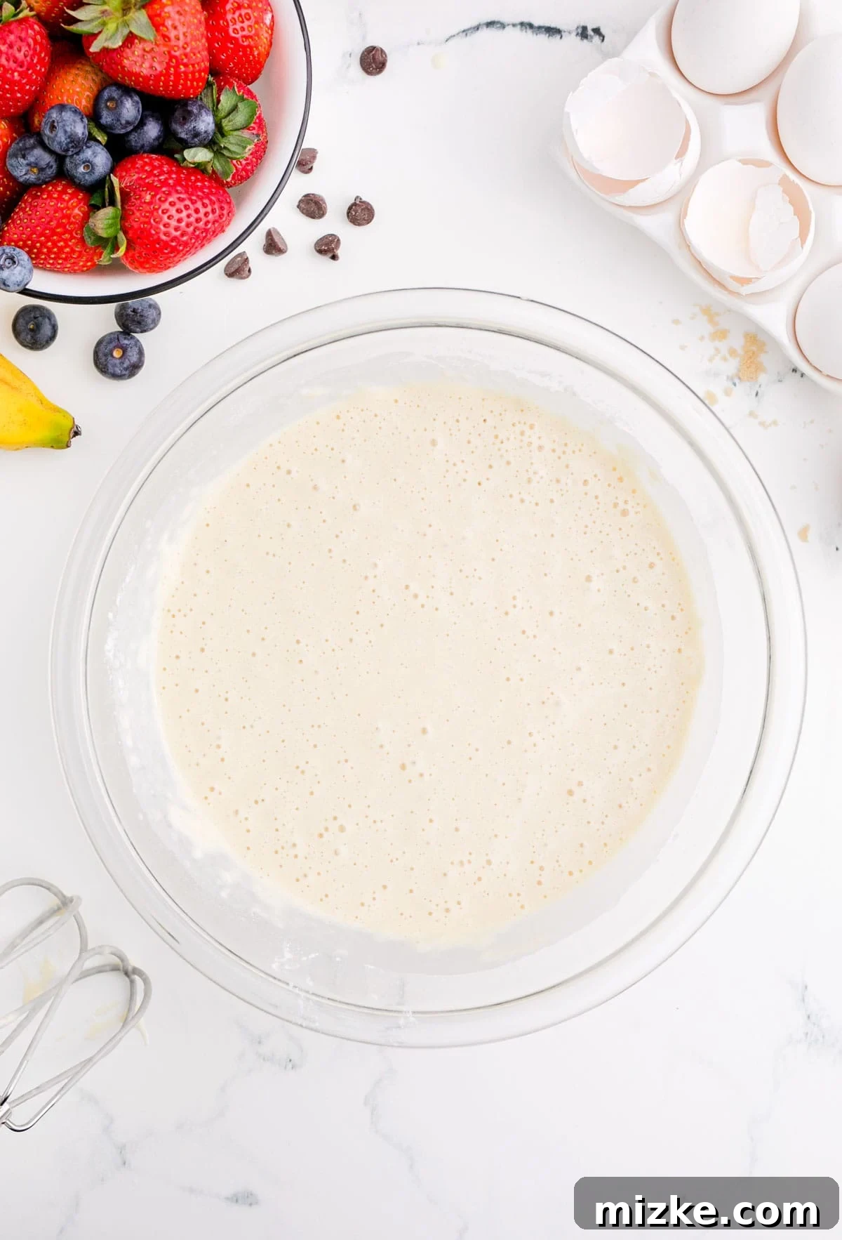One-Pan Fruity Pancakes 9 A bowl of smooth fruit pancake batter resting next to broken eggshells and fresh berries, indicating a ready-to-use mixture.
