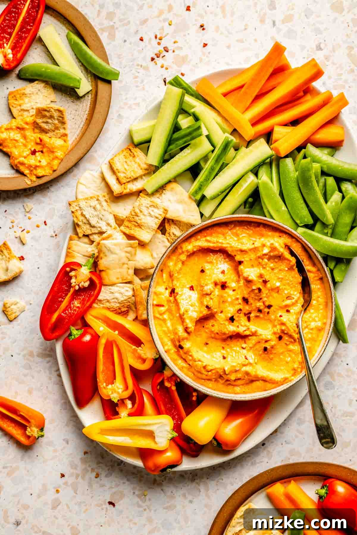 A platter with sliced vegetables, including carrots, cucumbers, and bell peppers, surround a bowl of kopanisti sprinkled with red pepper flakes, alongside pita chips.