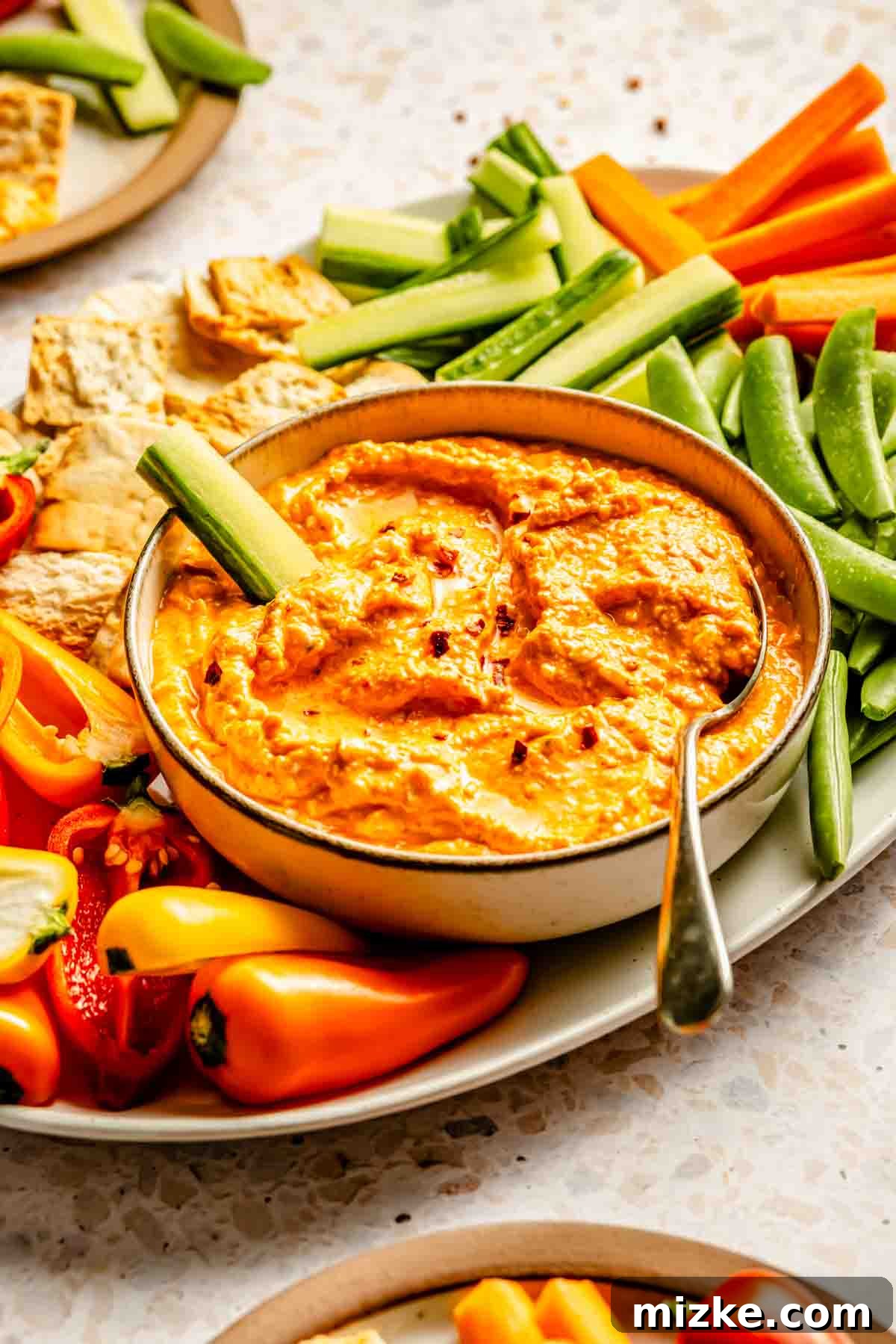 A bowl of kopanisti with a spoon, surrounded by sliced carrots, cucumbers, bell peppers, snap peas, and pita chips on a plate.