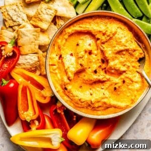 A bowl of kopanisti with a spoon, surrounded by sliced peppers, cucumbers, snap peas, pita slices, and crackers on a plate.