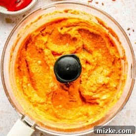 A food processor bowl filled with red puree, surrounded by garlic, a bowl of red bell peppers, and a small bowl of red pepper flakes on a light countertop.