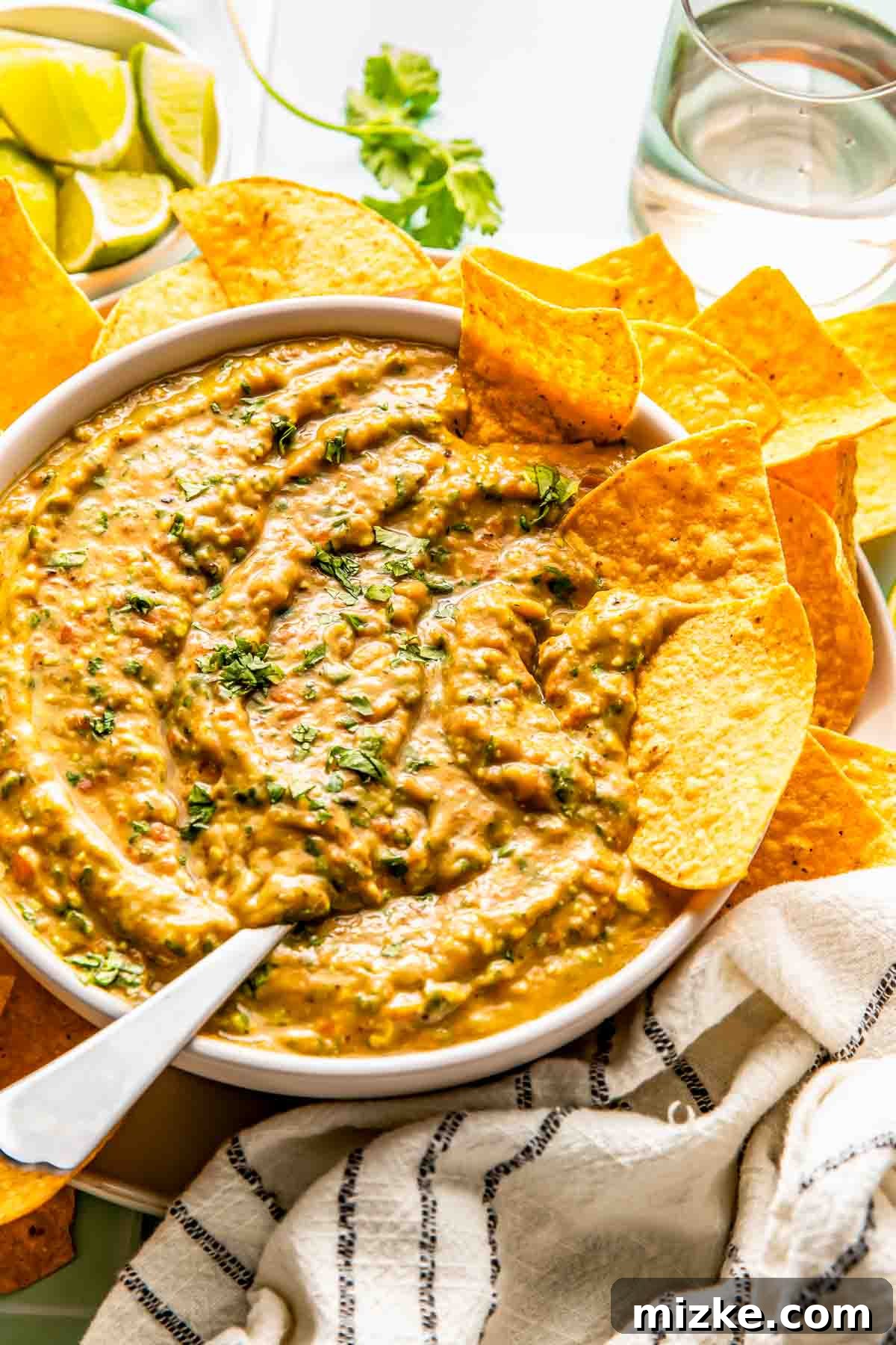 Teedos Legendary Salsa 3 A vibrant bowl of Teedo's creamy avocado salsa, garnished with fresh green herbs, is surrounded by a generous pile of golden tortilla chips. In the background, bright lime wedges and a refreshing glass of water hint at the perfect pairing for this delicious dip.