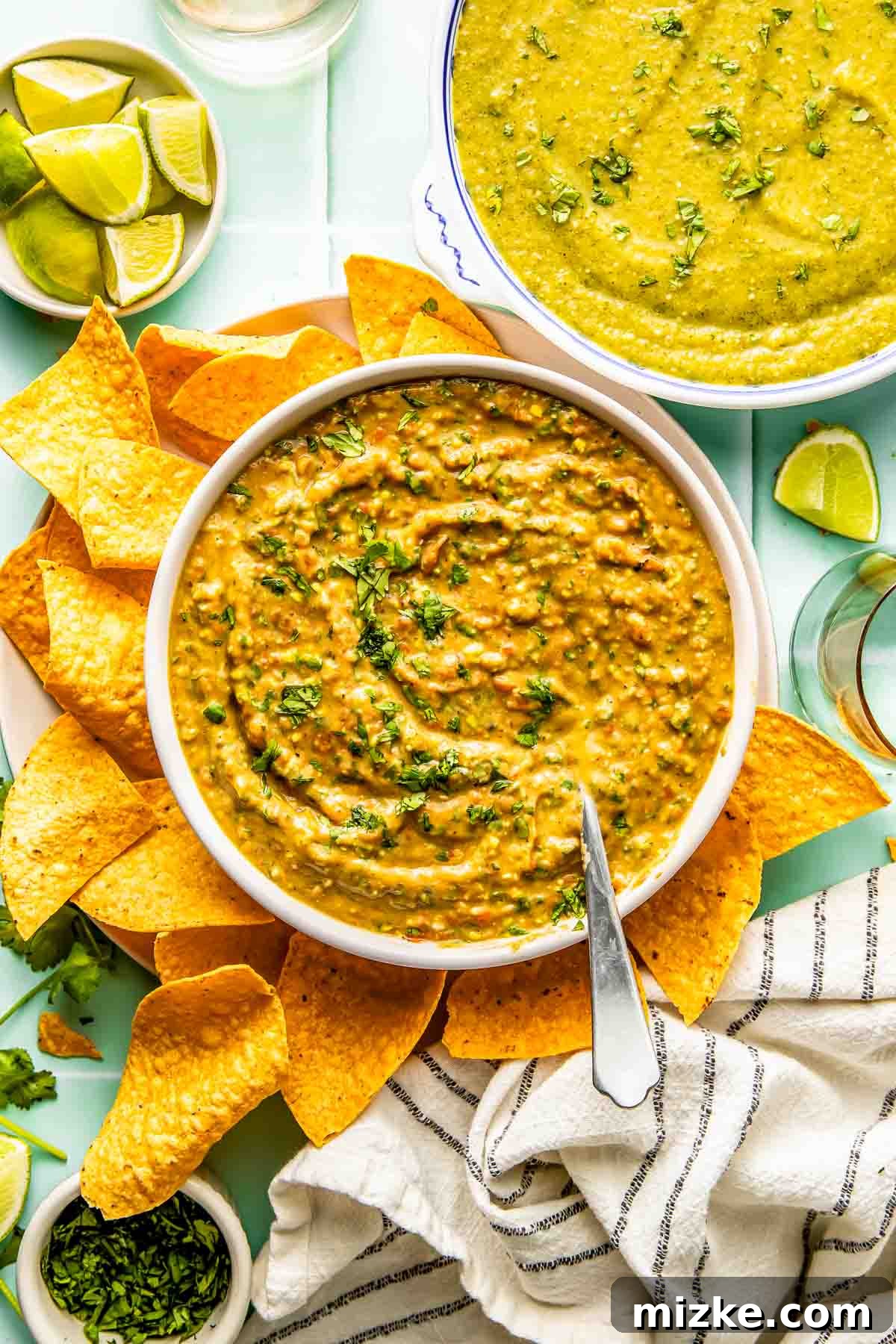 Teedos Legendary Salsa 4 A generously filled bowl of creamy Teedo's Famous Salsa, garnished with vibrant green herbs, takes center stage. It's surrounded by a plentiful scattering of golden-brown tortilla chips, inviting immediate indulgence. In the background, bright green lime wedges offer a zesty counterpoint, and another small bowl holds a different green dip, suggesting a delightful variety for a party spread.