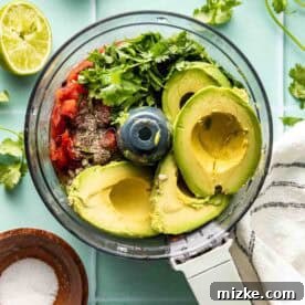 Teedos Legendary Salsa 6 Food processor filled with avocado, tomatoes, cilantro, spices, and lime halves nearby, on a light blue surface with a striped towel and a small bowl of salt.