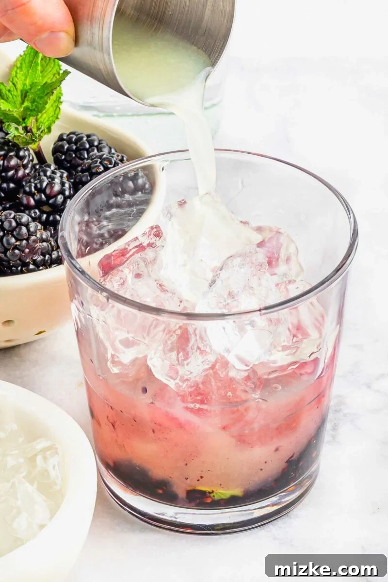 Velvet Blackberry Mint Mojito 5 Lime juice is being poured into a glass with muddled berries and mint at the bottom of it.