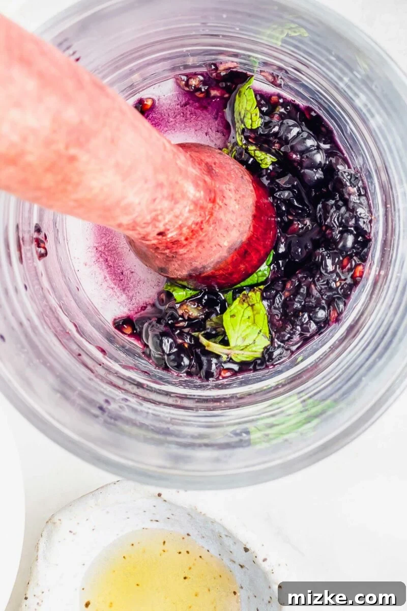 Velvet Blackberry Mint Mojito 6 A muddler is smashing berries and mint at the bottom of a glass.