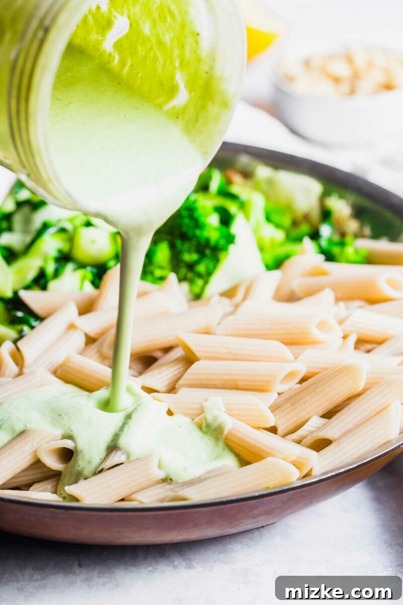 Velvet Cashew Basil Vegan Pasta 3 A vibrant green cashew basil pasta sauce is being generously poured over a bowl of freshly cooked pasta and sautéed vegetables.
