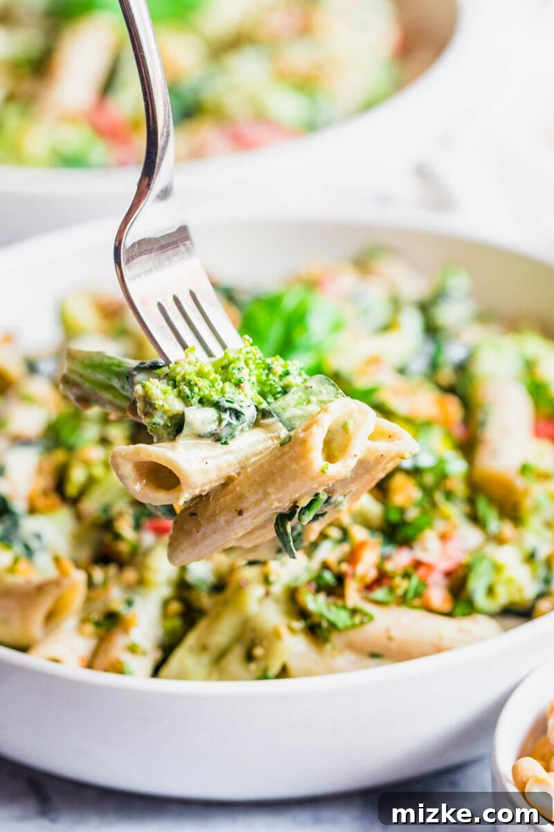 Velvet Cashew Basil Vegan Pasta 5 A fork lifts a generous serving of creamy vegan basil cashew pasta from a white bowl, showcasing the rich sauce and vibrant vegetables.