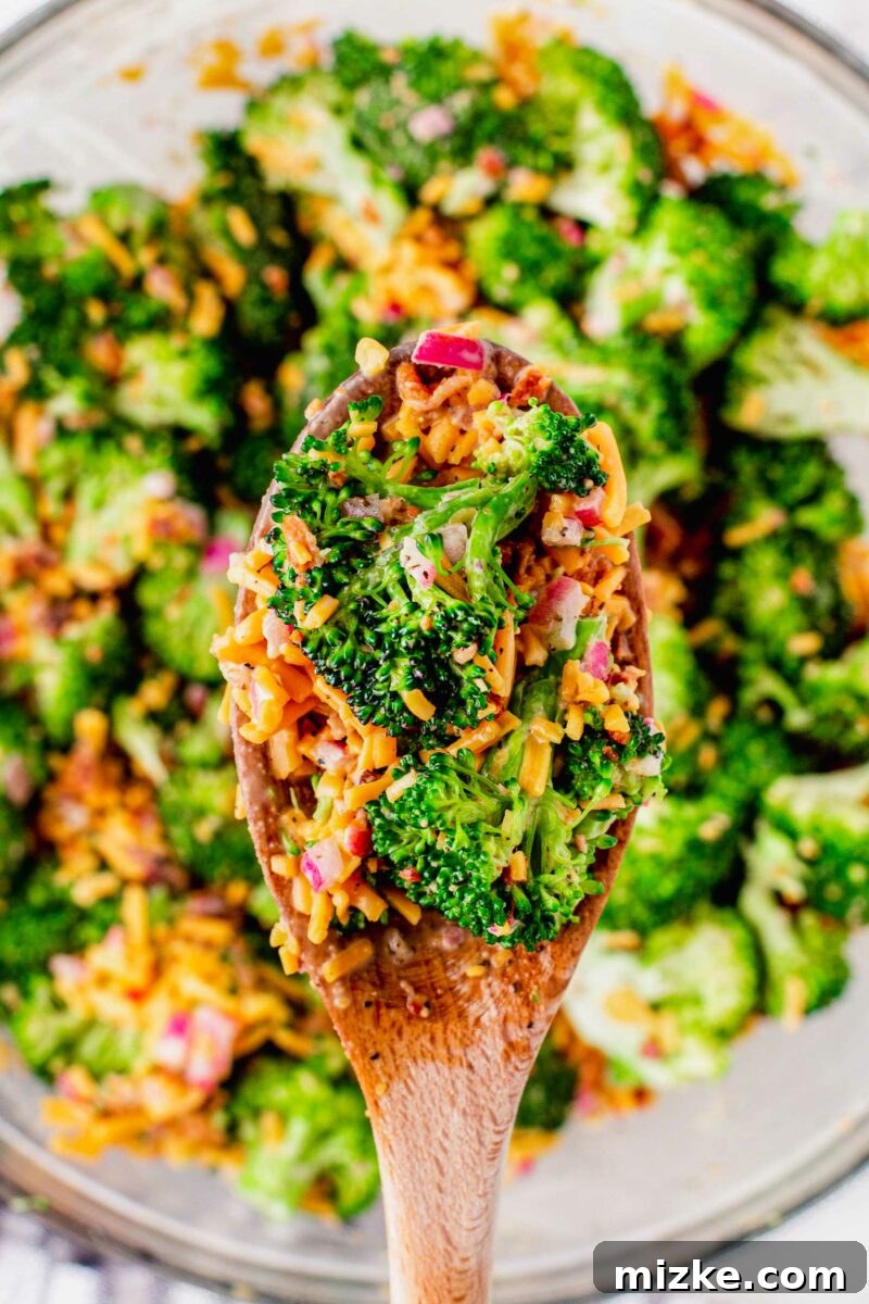 Crisp Broccoli Delight 2 A wooden spoon gently lifts a generous portion of vibrant broccoli salad from a large white serving bowl, showcasing the fresh green broccoli florets, bright orange shredded cheddar cheese, and savory bits of crumbled bacon, all coated in a creamy dressing.