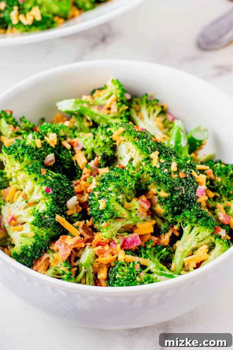 Crisp Broccoli Delight 5 A pristine white serving bowl is beautifully filled with freshly made creamy broccoli salad, garnished generously with extra shredded cheese and crispy bacon. The salad appears rich in texture and inviting, ready to be enjoyed as a centerpiece or a hearty side.