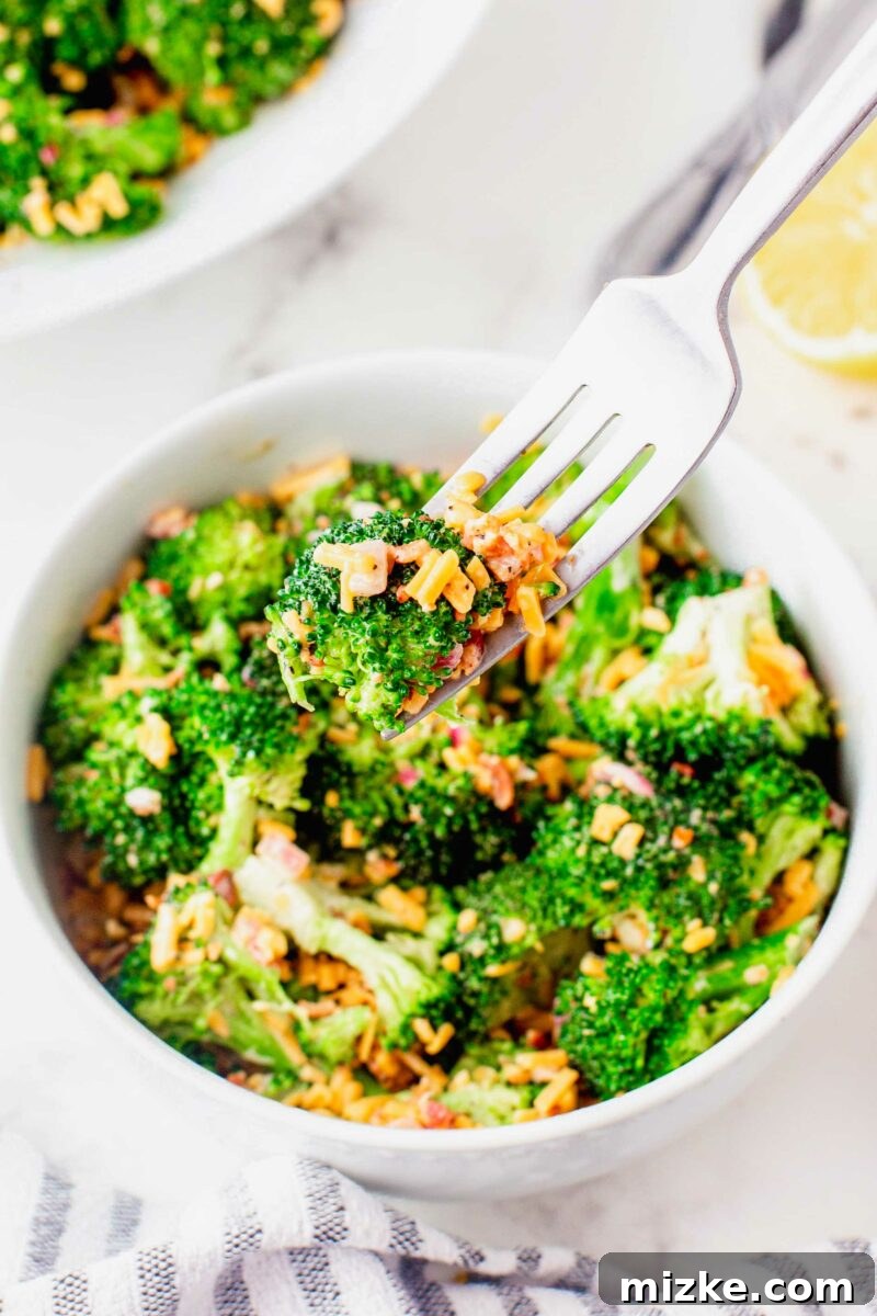Crisp Broccoli Delight 6 A fork has perfectly pierced a single, crisp piece of fresh broccoli, holding it aloft just above a generously filled bowl of creamy broccoli salad. This image highlights the inviting texture of the broccoli and signals that the delicious salad is ready to be savored.