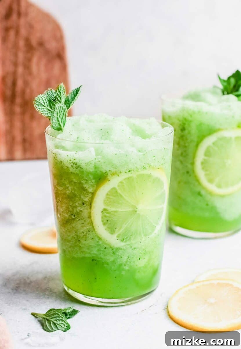 Two glasses of vibrant green frozen mint lemonade sit on a bright white countertop, garnished with fresh mint sprigs.