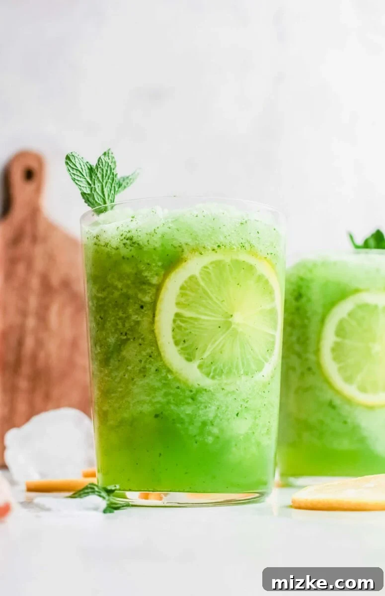 A vibrant green frozen lemonade, garnished with a lemon slice and mint sprig, is served in a clear glass on a rustic wooden surface.