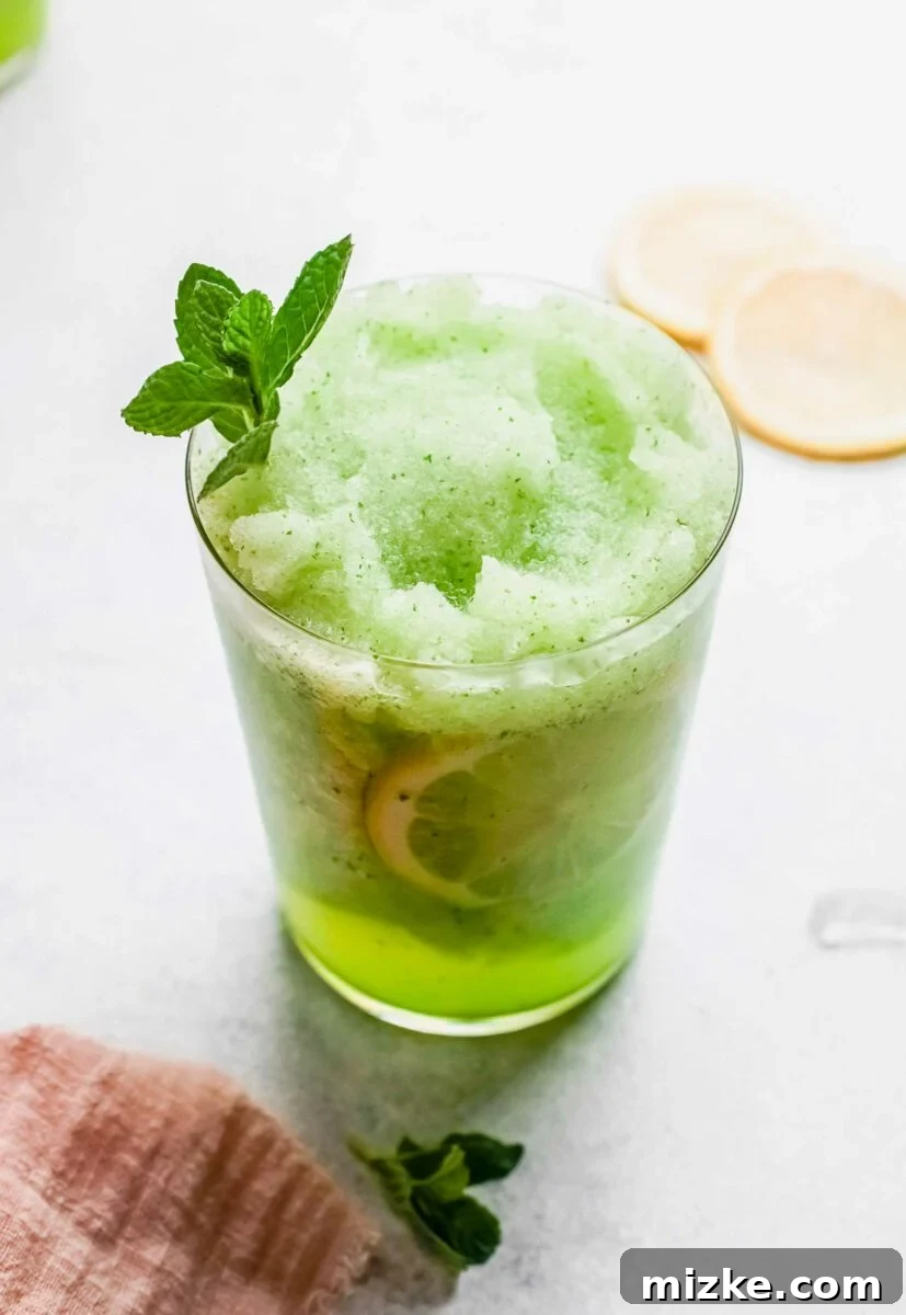 A single glass of frozen mint lemonade, garnished with a mint sprig, is presented on a clean white countertop, highlighting its refreshing appeal.