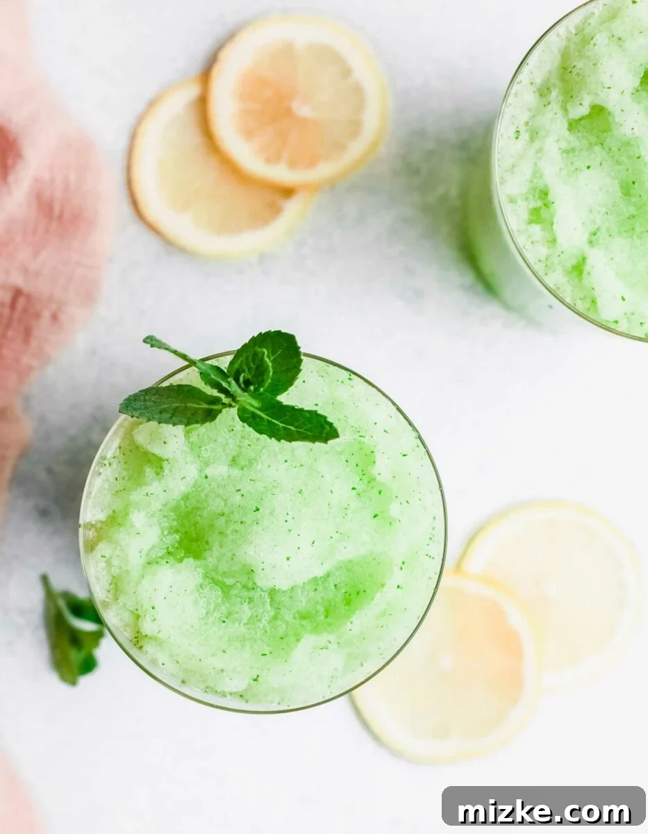 Lemon slices and a sprig of mint are artfully arranged next to a glass of frozen mint lemonade, emphasizing the fresh ingredients.