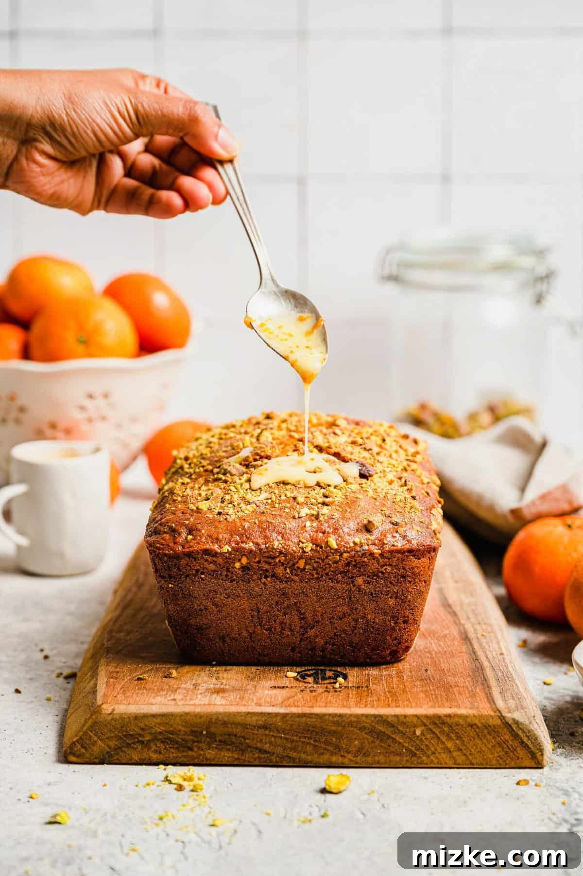Zesty Orange Pistachio Loaf 6 A hand drizzling the vibrant orange glaze over a baked loaf of orange pistachio bread, with green pistachios visible.