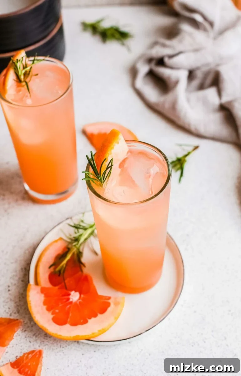Bitter Sweet Gin and Tonic 4 Two glasses of grapefruit gin and tonic are arranged on a white surface, accompanied by extra slices of fresh grapefruit and rosemary for garnish.