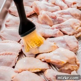raw chicken wings on a metal baking sheet being brushed with oil with a silicone brush