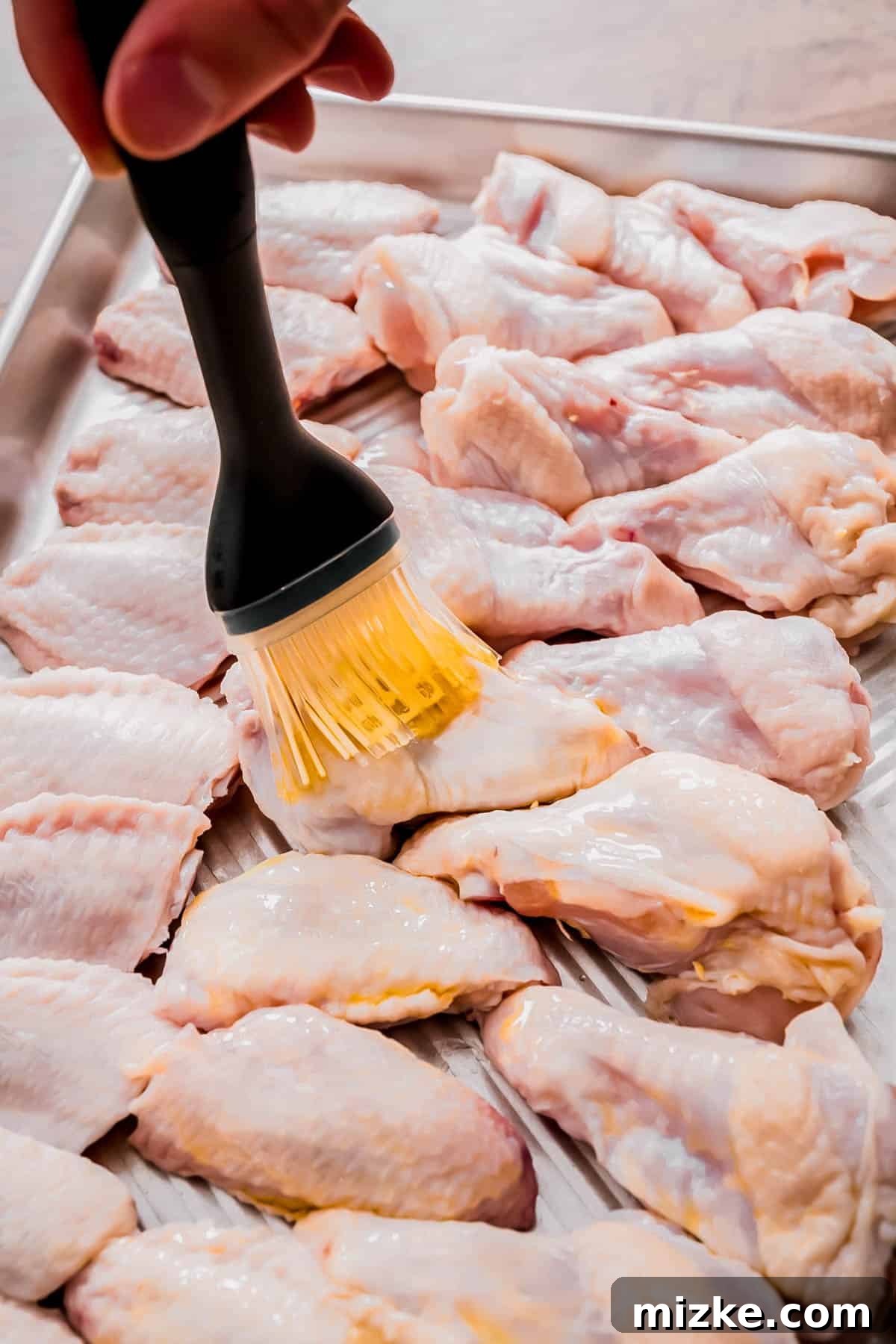 Irresistible Crispy Grilled Wings 4 Raw chicken wings arranged on a metal baking sheet, being lightly brushed with avocado oil using a silicone brush, prior to seasoning and grilling.