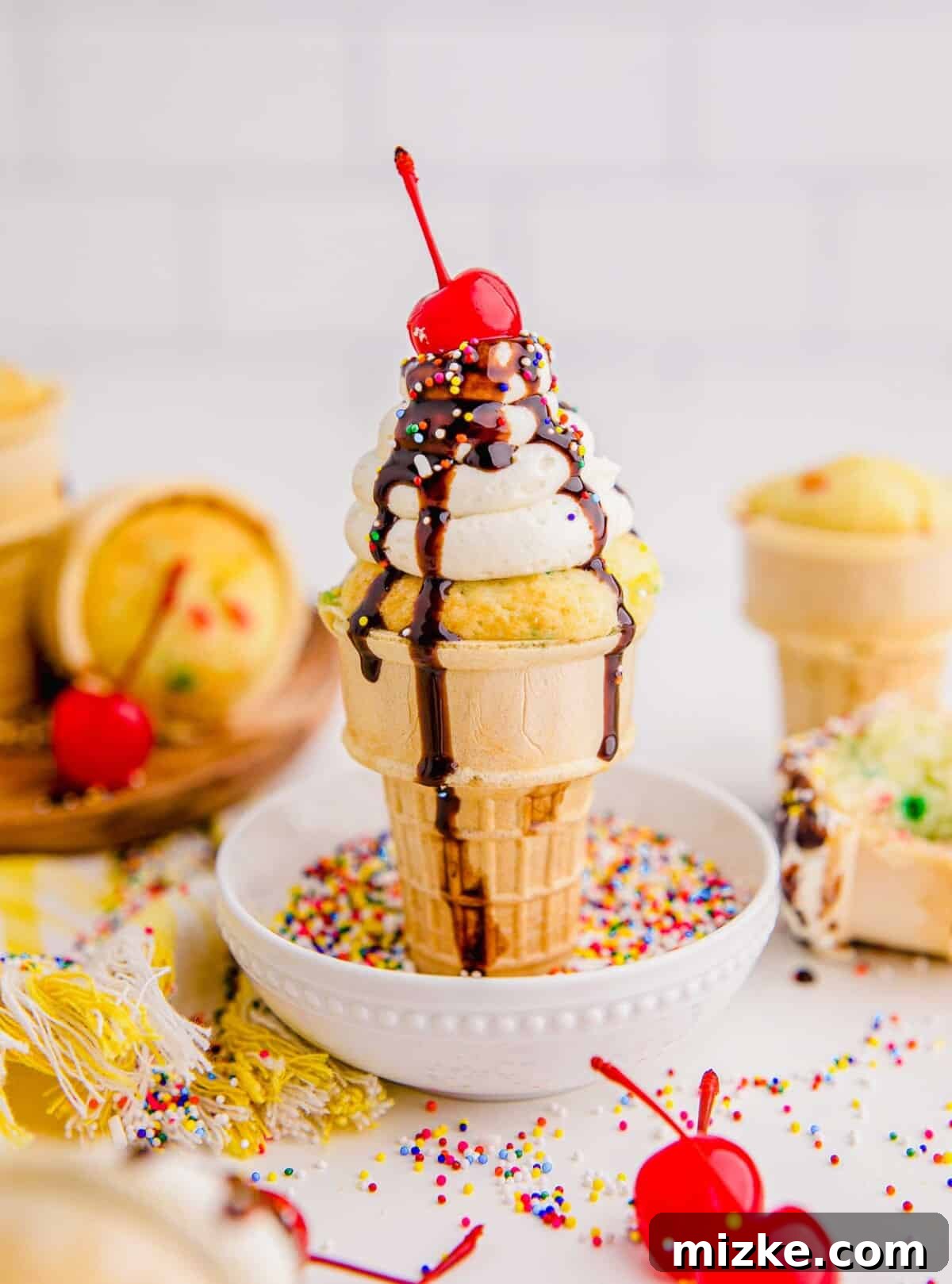 Waffle Cone Cupcakes 13 An ice cream cone cupcake stands upright in a small white bowl, which is filled with an abundance of rainbow sprinkles, ready for a fun garnish.
