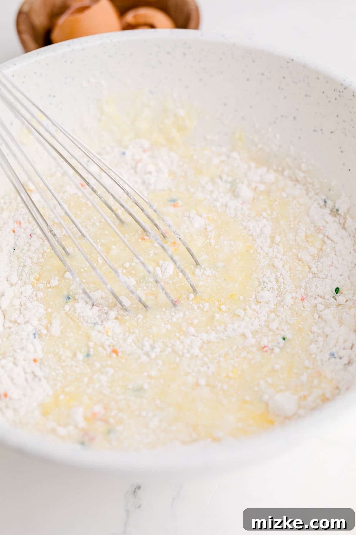 Waffle Cone Cupcakes 5 A metal whisk diligently mixing funfetti cake batter in a speckled mixing bowl.