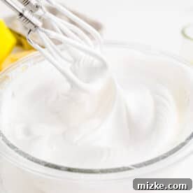 Fluffy white meringue topping is being whipped to perfection with an electric whisk in a clear bowl, ready to crown the cupcakes.