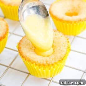 A spoonful of vibrant yellow lemon curd is being carefully placed into a cored-out lemon cupcake, creating a delicious surprise center.