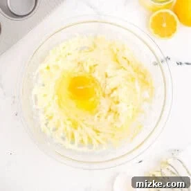 A cracked egg being added to the creamy butter and sugar mixture in a large mixing bowl, ready for blending.