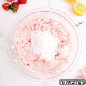 Additional powdered sugar is added to the pink strawberry frosting mixture in a clear bowl for desired consistency