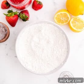 Dry ingredients in a white bowl next to a metal whisk and fresh strawberries and lemons