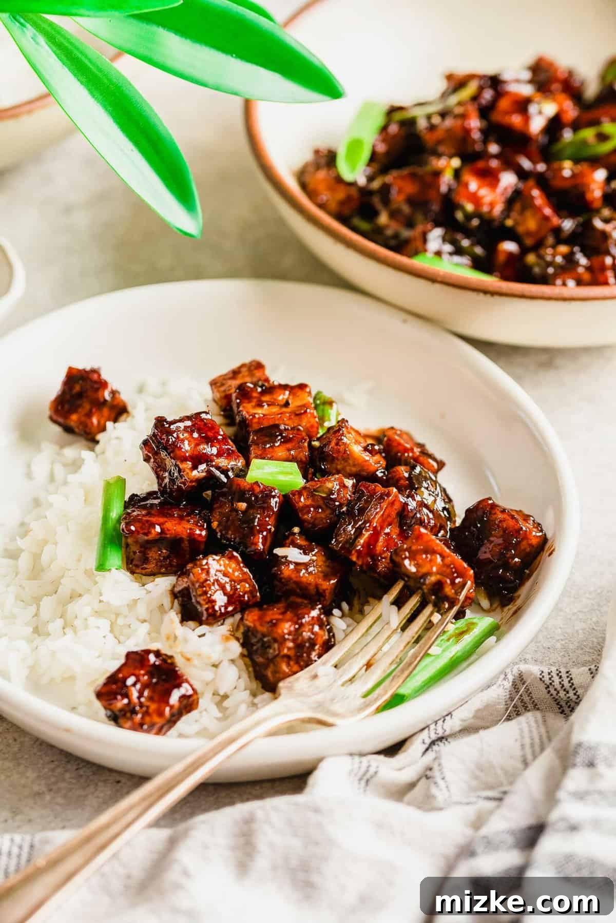 This black pepper tofu dish is a meatless entree that is sure to satisfy! The sauce has bold flavors that coat extra crispy tofu cubes! A fork is poised over a white bowl filled with Black Pepper Tofu, fluffy rice, and fresh spring onions, ready for a delicious bite.